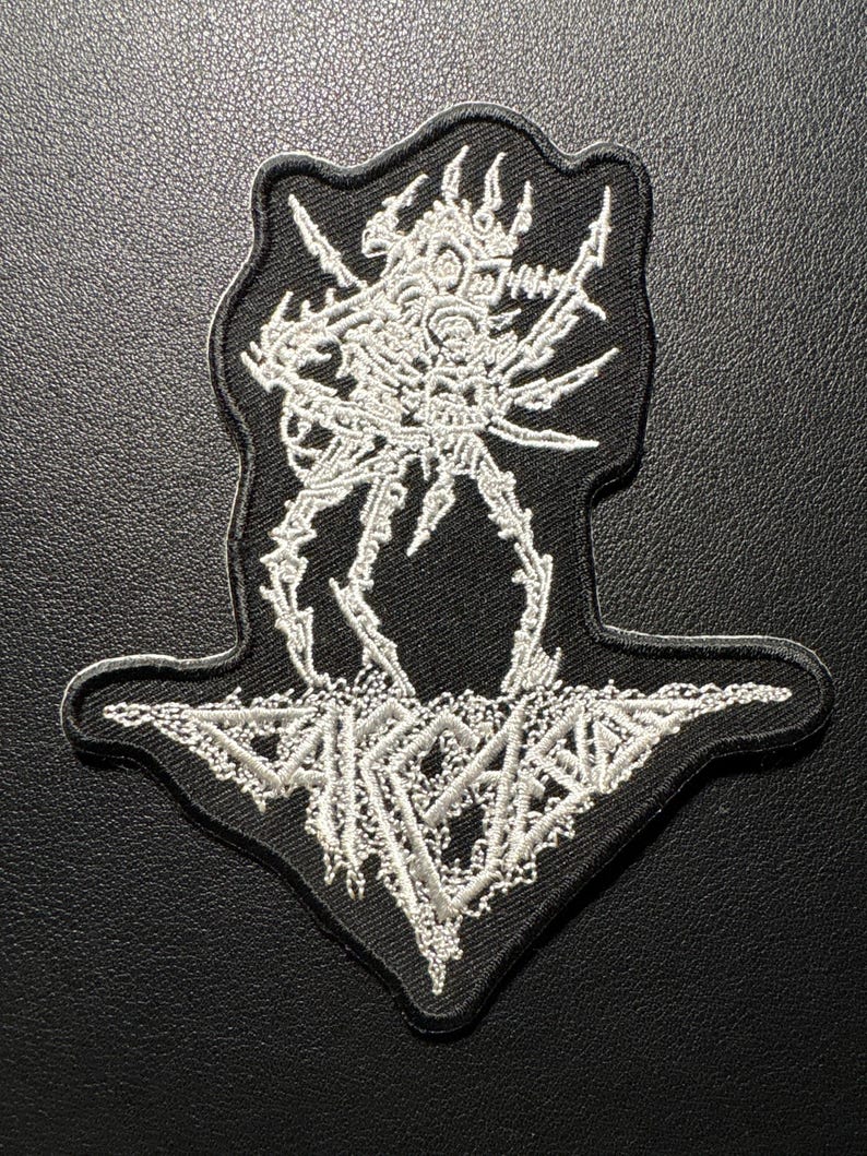 以下が含まれることがあります： 白黒の刺繍パッチで、骸骨のような生き物のデザインが特徴です。 パッチはユニークな形で、骸骨の姿と「SARCOMA」という言葉が白で詳細に描かれています。