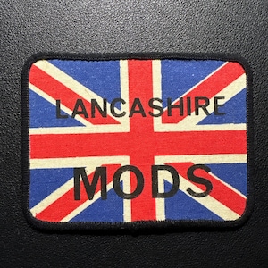 May include: A rectangular patch featuring the Union Jack flag design with the words "LANCASHIRE MODS" in black block letters. The patch has a black border and is set against a dark background.