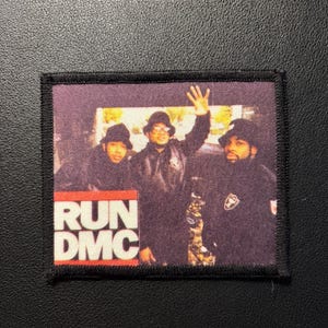 Op de afbeelding: Een vierkante stoffen patch met een gedrukte foto van de hiphopgroep Run-DMC. De drie leden dragen zwarte bucket hats en donkere jassen. De tekst RUN DMC is in witte hoofdletters gedrukt op een rode rechthoekige achtergrond.