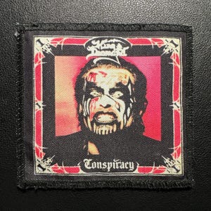 Può includere: Una toppa rettangolare con un ritratto di una persona con trucco bianco e nero e dettagli rossi. La toppa ha un bordo nero e la parola "Conspiracy" in basso.