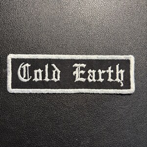 Könnte beinhalten: Ein rechteckiger schwarzer Aufnäher mit den Worten "Cold Earth" in weißer gotischer Schrift gestickt. Der Aufnäher hat einen weißen Rand und ist vor einem strukturierten schwarzen Hintergrund platziert.
