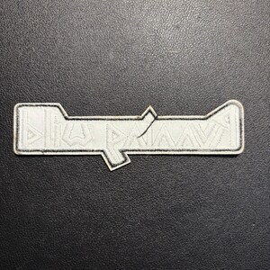 May include: A white embroidered patch with a geometric design and black border. The patch has a rectangular shape with a stylized angular design. The patch is on a black textured surface.