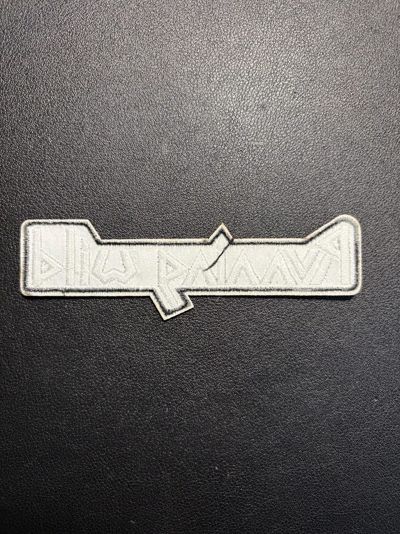 May include: A white embroidered patch with a geometric design and black border. The patch has a rectangular shape with a stylized angular design. The patch is on a black textured surface.