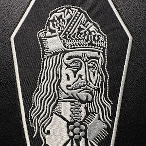 Può includere: Una toppa ricamata nera a forma di bara, con un ritratto dettagliato di un uomo con corona e capelli lunghi. Il ritratto è in filo bianco con dettagli intricati, delineato da un bordo bianco.