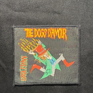 May include: A rectangular patch featuring the text "THE DOGS D'AMOUR" in orange and a cartoon figure in a green coat and red hat. The figure is holding a flaming object. The text "EARL FRY" is on the left side.