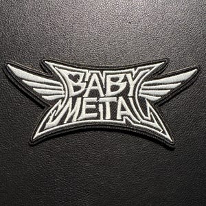 Könnte beinhalten: Schwarz-weißer, gestickter Aufnäher mit dem Schriftzug "BABY METAL" in einem stilisierten Schriftzug. Das Design beinhaltet Flügel. Geeignet für Kleidung oder Accessoires.