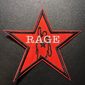 Könnte beinhalten: Ein roter sternförmiger Aufnäher mit schwarzem Rand und dem Wort "RAGE" in weißen Blockbuchstaben. Eine Silhouette einer Person mit erhobenem Arm befindet sich in der Mitte.
