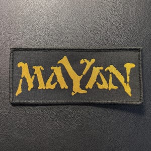 Puede incluir: Un parche rectangular bordado con fondo negro y la palabra MAYAN cosida en hilo amarillo dorado texturizado. Las letras tienen un estilo irregular y desgastado, y todo el parche está rematado con un borde negro cosido alrededor.
