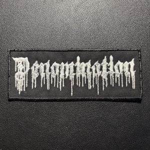May include: A rectangular black fabric patch featuring white embroidered text that reads "Denomination" in a stylized, dripping font. The embroidery has a textured appearance with vertical lines extending downward from the letters, creating a melting effect.