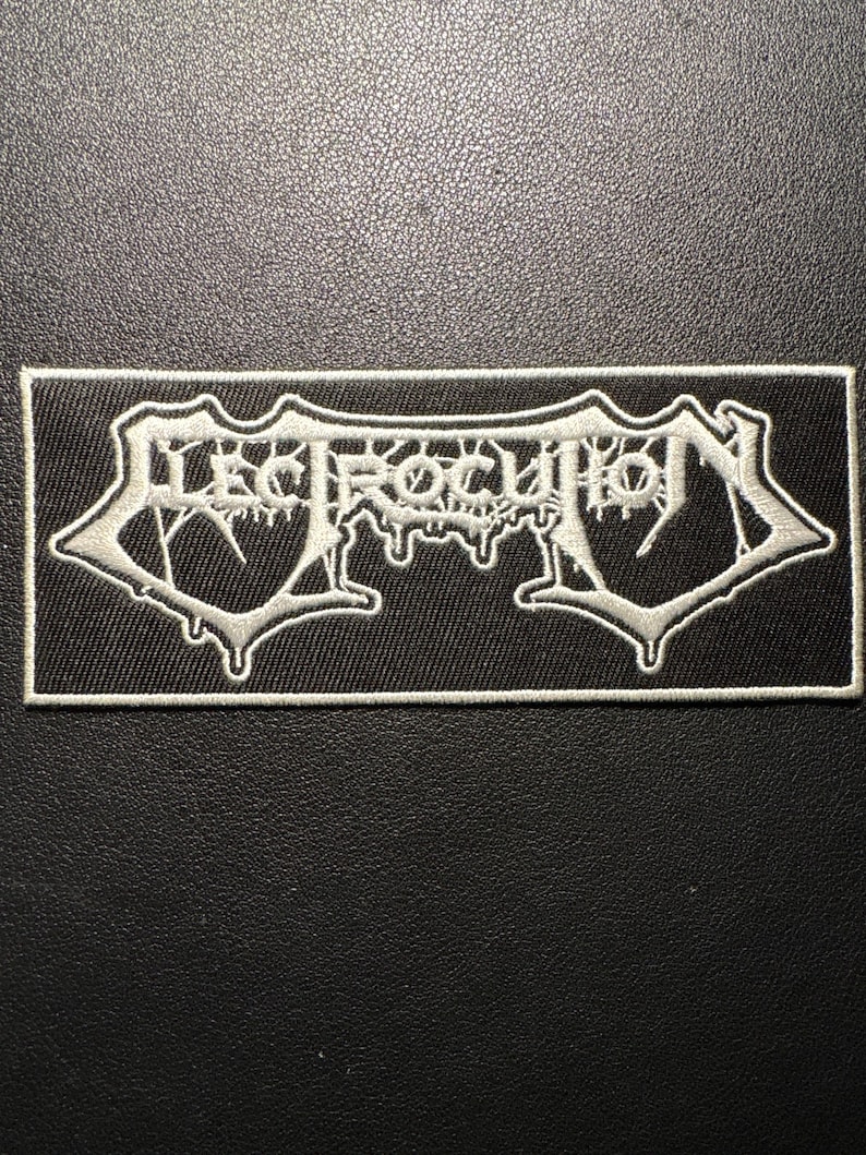 May include: A black rectangular patch featuring the word "ELECTROCUTION" in white, gothic-style lettering. The patch has a white border. Likely designed for application on clothing or accessories.
