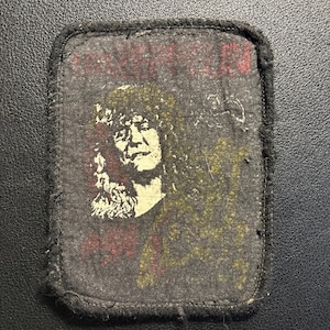 Könnte beinhalten: Ein rechteckiger schwarzer Stoffaufnäher mit abgerundeten Ecken, der eine Siebdruckgrafik einer Person mit langem Haar in einem hellgelben Ton zeigt. Der Text Led Zeppelin ist oben in Rot gedruckt und eine gelbe Gitarrenform ist sichtbar.