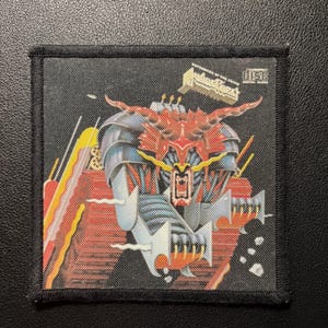 May include: A square fabric patch with a black border featuring a graphic illustration of a red and blue mechanical creature with sharp horns and claws. The design includes yellow accents and a small compact disc logo in the upper right corner.