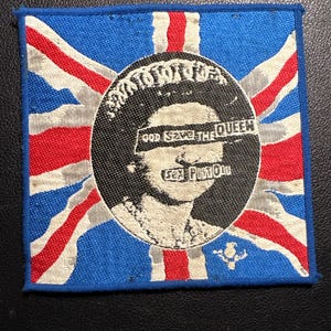 Könnte beinhalten: Quadratischer Aufnäher mit Union Jack-Design und einem zentralen kreisförmigen Bild. Das Bild zeigt eine Person mit dem Text "GOD SAVE THE QUEEN" und "SEX PISTOLS". Der Aufnäher hat einen blauen Rand.