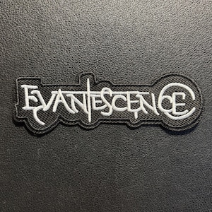 May include: A black embroidered patch featuring the word EVANESCENCE in white stylized lettering. The text is outlined in white thread against a black fabric background, creating a high-contrast design with a distinct, jagged font style.