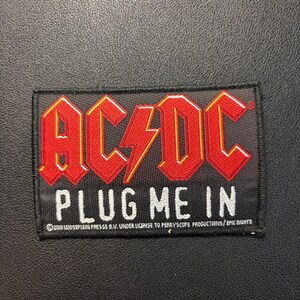 May include: A rectangular embroidered patch with a black background. The red AC/DC logo is at the top with a yellow lightning bolt. Below, the white text reads PLUG ME IN. Small white text at the bottom reads 2019 LEIDSEPLEIN PRESSE B.V. UNDER LICENSE TO PERRYSCOPE PRODUCTIONS/EPIC RIGHTS.