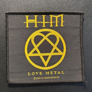 Könnte beinhalten: Ein quadratischer schwarzer Stoffaufnäher mit gelber Stickerei. Oben ist der Text HIM zu sehen, in der Mitte ein Heartagram-Symbol und unten der Text LOVE METAL mit dem darunter stehenden Copyright-Text 2004 OY HEARTAGRAM LTD.