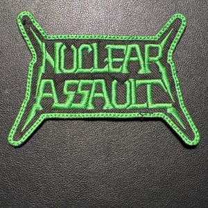 Könnte beinhalten: Gestickter Aufnäher mit den Worten "NUCLEAR ASSAULT" in leuchtend grüner Farbe. Der Text ist in einer kräftigen, stilisierten Schriftart gehalten, grün umrandet und vor einem schwarzen Hintergrund platziert. Der Aufnäher hat einen einzigartigen, eckigen Rand.