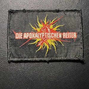 Può includere: Toppa rettangolare nera con la scritta "DIE APOKALYPTISCHEN REITER" in rosso. La toppa presenta un disegno centrale di un'esplosione gialla e rossa. La toppa è probabilmente per una giacca o una borsa.