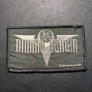 Puede incluir: Un parche rectangular negro con el nombre de la banda "Dimmu Borgir" en letras plateadas. El parche presenta un pentagrama y un diseño estilizado. También se ve el texto "NUCLEAR BLAST 2003".