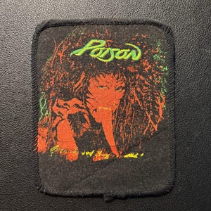 Pode incluir: Um emblema retangular de tecido preto com borda costurada, apresentando uma estampa gráfica de uma pessoa com cabelo volumoso. A palavra Poison está impressa em verde no topo, com detalhes em laranja e amarelo na ilustração central.