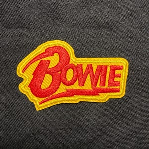 Op de afbeelding: Een geel en rood geborduurde patch met de naam "Bowie" in een gestileerd lettertype. De patch heeft een gele rand en is geplaatst op een zwarte stoffen achtergrond. Het woord "Bowie" is rood.