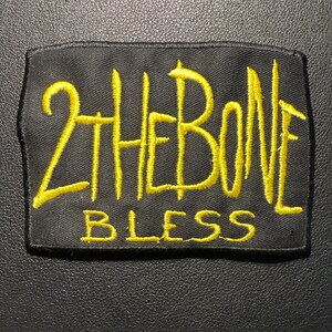 Könnte beinhalten: Schwarzer rechteckiger Aufnäher mit gelbem gesticktem Text. Der Text lautet "2 THE BONE BLESS". Der Aufnäher ist für Kleidung oder Accessoires bestimmt, mit einem einfachen, kräftigen Design.