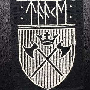 Könnte beinhalten: Schwarzer rechteckiger Aufnäher mit weißem Emblem. Das Emblem zeigt ein Schilddesign mit vertikalen Linien, gekreuzten Äxten und einer Krone. Oberhalb des Schildes befinden sich stilisierte weiße Buchstaben und Punkte.