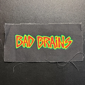 May include: A rectangular black fabric patch featuring the text BAD BRAINS in a stylized font. The letters are colored red with a yellow outline and a green border, creating a vibrant contrast against the dark, textured fabric background.