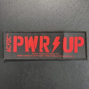 May include: A rectangular black embroidered patch featuring the red text PWR/UP with a lightning bolt symbol in place of the slash. The AC/DC logo is on the left, and small white text at the bottom reads 2020 LEIDSEPLEIN PRESSE B.V. UNDER LICENSE TO PERRYSCOPE PRODUCTIONS, LLC/EPIC RIGHTS, LLC.