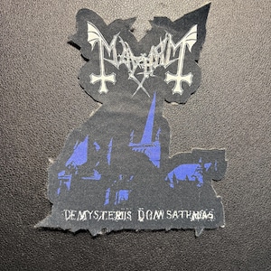May include: A black patch featuring the band name "Mayhem" in white gothic lettering, with inverted crosses and a bat-like design. Below, a blue and black graphic depicts a silhouette of a building, with the text "De Mysteriis Dom Sathanas".