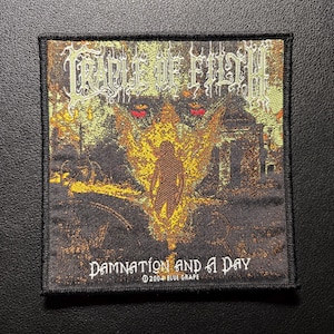 Op de afbeelding: Vierkantige patch met de bandnaam "Cradle of Filth" in sierlijke letters boven een illustratie van een figuur in vlammen. De tekst "Damnation and a Day" staat eronder, met "© 2004 BLUE GRAPE" eronder. De patch heeft een zwarte rand.