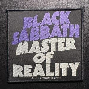 Puede incluir: Parche de Black Sabbath "Master of Reality". El parche rectangular presenta el nombre de la banda en morado y el título del álbum en blanco. El parche está sobre un fondo negro con un borde negro. La fecha de copyright es 2013.