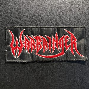 Könnte beinhalten: Ein rechteckiger, bestickter Aufnäher mit schwarzem Hintergrund und dem Wort Warbringer, das in einer stilisierten, scharfkantigen roten Schrift gestickt ist. Die Schrift hat eine aggressive Ästhetik mit spitzen Serifen.