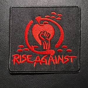 Könnte beinhalten: Schwarzer quadratischer Aufnäher mit rotem gesticktem Design und Text. Das Design zeigt eine geballte Faust in einem Kreis mit einer stilisierten Flamme und den Worten "RISE AGAINST" in fetter Schrift. Der Aufnäher ist wahrscheinlich für Kleidung oder Accessoires.