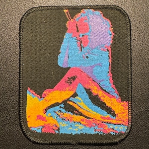 Könnte beinhalten: Ein schwarzer, rechteckiger Aufnäher mit einer farbenfrohen, stilisierten Illustration einer Person, die ein Saiteninstrument spielt. Die Figur ist in Blau-, Rosa-, Orange- und Lilatönen dargestellt. Der Aufnäher hat einen genähten Rand.