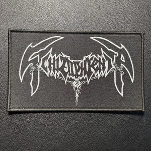 May include: A rectangular black embroidered patch featuring white thread detailing. The design displays stylized, sharp-edged lettering that reads Schizophrenia, accented with chain-like elements and a central eye graphic hanging from the text.