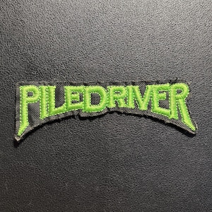 Può includere: Una toppa in tessuto nero con la parola PILEDRIVER ricamata in filo verde brillante. Il testo è stilizzato con una leggera curva e lettere in grassetto, con un bordo nero che delinea il ricamo verde sulla base in tessuto.