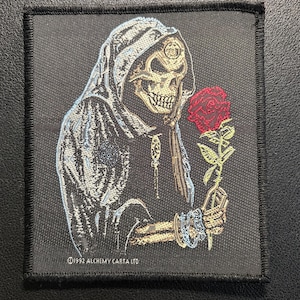May include: A square black embroidered patch featuring a hooded skeleton figure wearing a robe and holding a red rose with green leaves. The skeleton has a gold-toned skull and wears bracelets. The text 1992 ALCHEMY CARTA LTD is printed in white at the bottom.