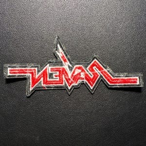 以下が含まれることがあります： 赤色の「SEVEN」という文字が白で縁取られ、ギザギザとした角張ったデザインの長方形パッチ。文字は様式化されています。パッチは黒い表面にあります。
