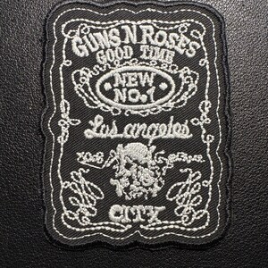 Könnte beinhalten: Schwarzer, gestickter Aufnäher mit weißen Nähten. Der Aufnäher zeigt den Text "GUNS N ROSES GOOD TIME NEW NO.1 Los angeles CITY" und ein Totenkopf-Design mit Rosen. Der Aufnäher hat einen dekorativen Rand.