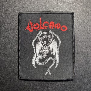 May include: Black square patch with a red and white design. The word "Volcano" is in red, stylized lettering above a white illustration of a winged creature with a serpent tail. The patch has a black border.