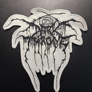 May include: A white embroidered patch featuring the black text Darkthrone in a stylized, dripping, illegible font. The patch has an irregular, organic shape with pointed edges and a thick white border, showcasing detailed black thread embroidery.