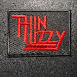 Könnte beinhalten: Ein rechteckiger, schwarzer Aufnäher mit dem Namen "THIN LIZZY" in roter Schrift. Der Schriftzug ist in einem speziellen Stil gehalten. Der Aufnäher ist zum Aufnähen auf Kleidung oder Accessoires gedacht.
