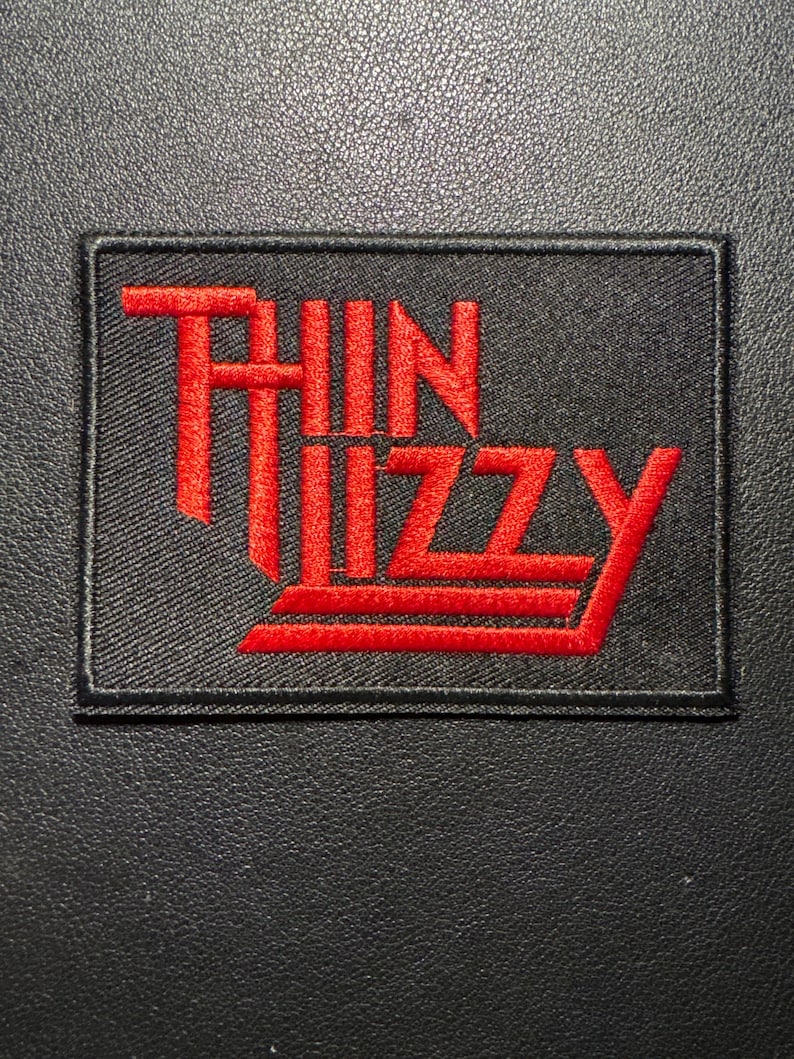 Pode incluir: Um patch retangular preto com o nome "THIN LIZZY" bordado em vermelho. O texto est&aacute; em uma fonte estilizada. O patch &eacute; projetado para ser costurado em roupas ou acess&oacute;rios.