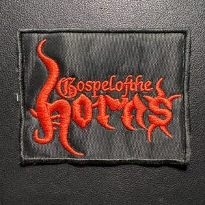 Peut inclure: Un écusson rectangulaire noir avec le texte brodé en rouge "Gospel of the Horns" dans une police gothique. L'écusson a une bordure noire et est probablement en tissu. Le fond est une surface sombre et texturée.