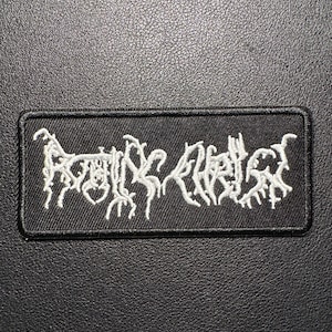 May include: A rectangular black patch with white embroidered text. The text is stylized and appears to be a band name or logo. The patch has a black border and is likely made of fabric.