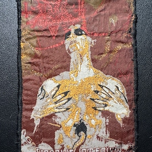 Könnte beinhalten: Gestickter Aufnäher mit dunkelrotem Hintergrund und schwarzem Rand. Der Aufnäher zeigt eine Figur mit goldenen und weißen Details und den Text "DIABOLIS INTERI" in Weiß. Die Oberseite des Aufnähers hat roten Text und ein Symbol.