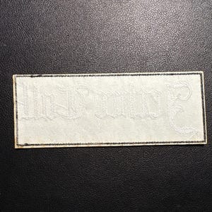 May include: A rectangular white patch with the words "In Dulce" embroidered in a decorative, gothic-style font. The patch has a black border and is set against a dark background.