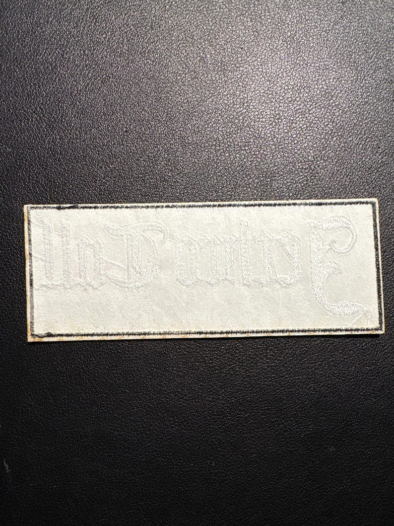 May include: A rectangular white patch with the words "In Dulce" embroidered in a decorative, gothic-style font. The patch has a black border and is set against a dark background.