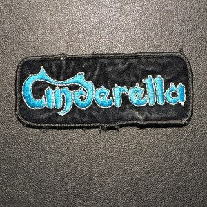 Pode incluir: Patch retangular preto com a palavra "Cinderella" bordada em turquesa. O patch tem um fundo preto aveludado e uma borda costurada. O texto está em uma fonte estilizada.